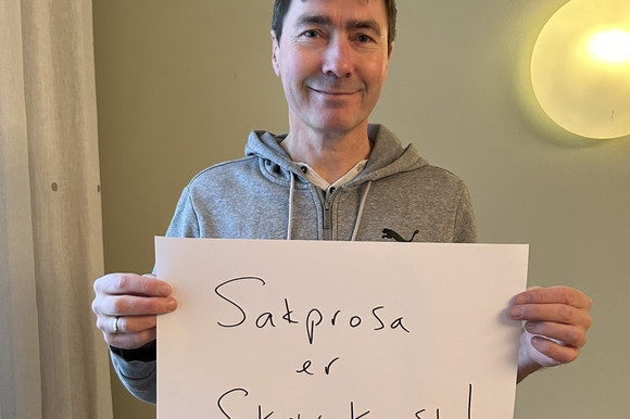 Mann med mørkt, kort hår som holder en plakat med skriften "Sakprosa er skrivekunst!".