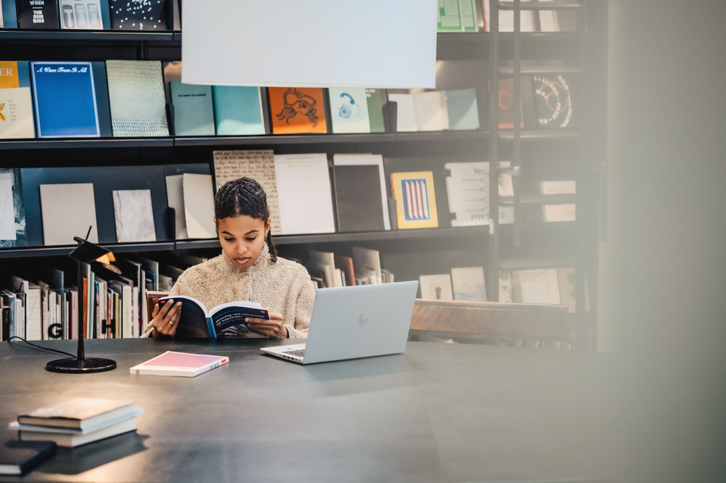 Kvinnelig student som leser i bok på bibliotek. Hun sitter ved et større bord og har en laptop slått opp foran seg. I bakgrunnen er hyller med utstilte bøker.