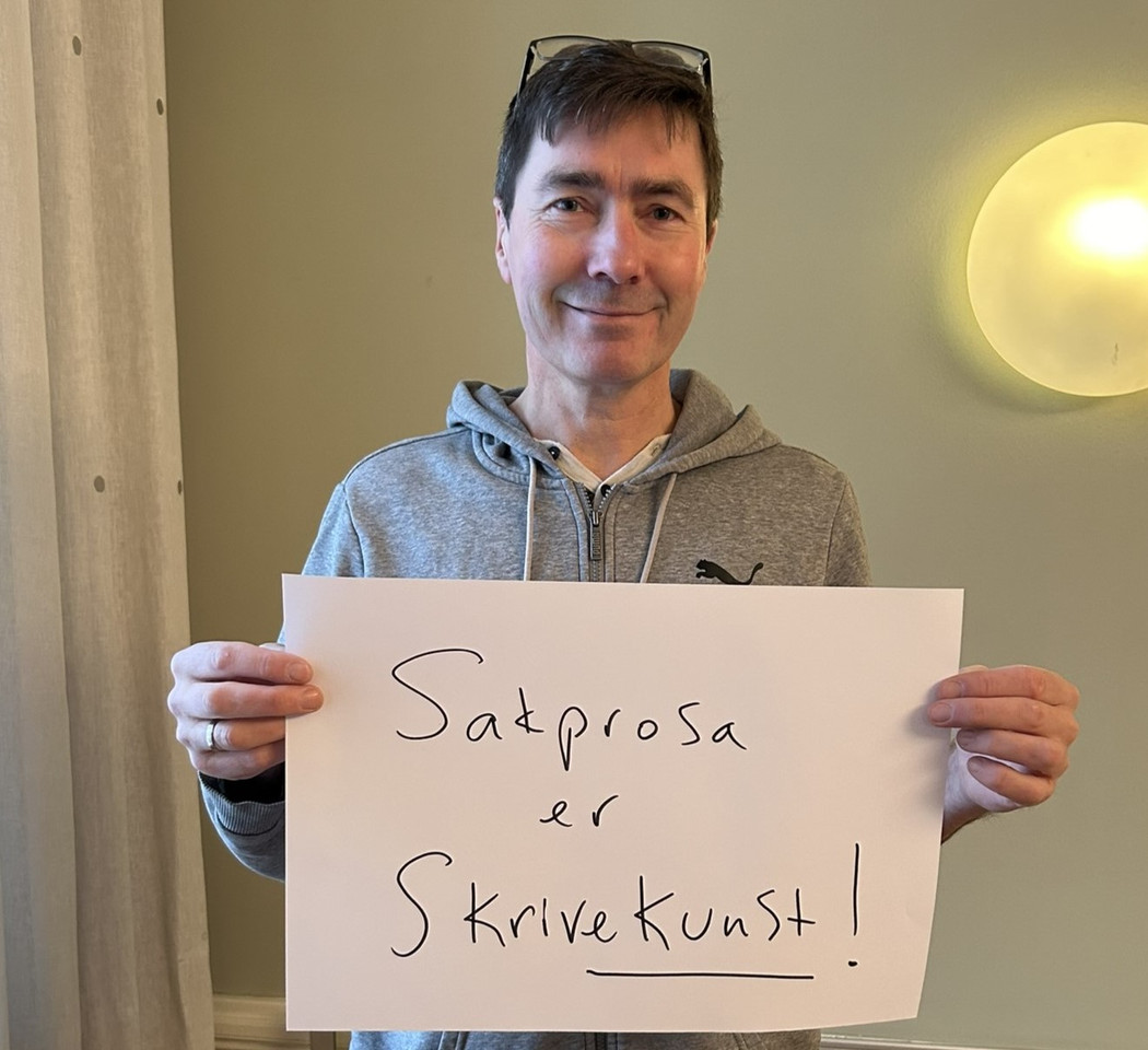 Mann med mørkt, kort hår som holder en plakat med skriften "Sakprosa er skrivekunst!".