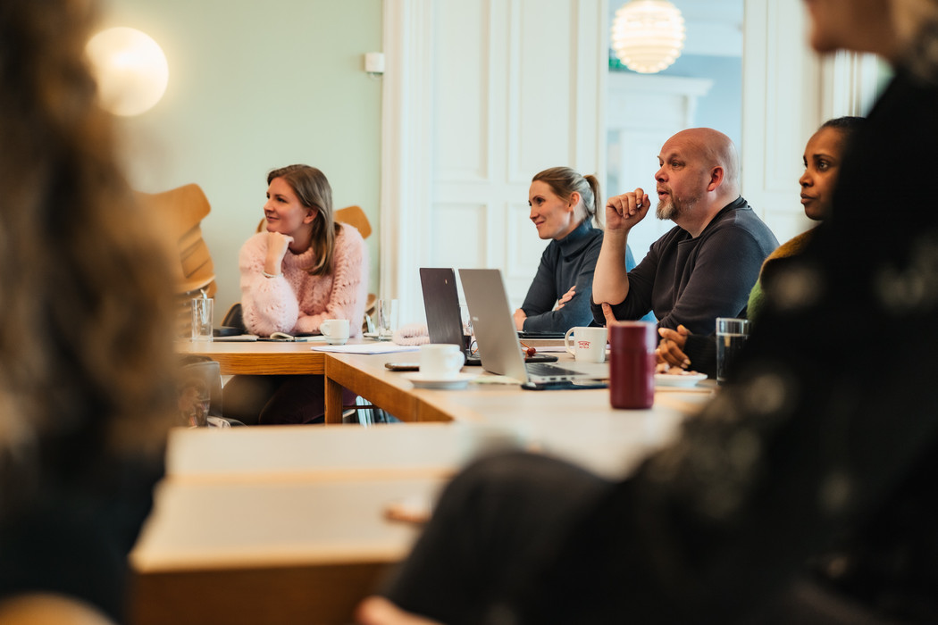 Voksne mennesker sitter ved skrivebord, blikket deres er vendt i samme retning, de følger med.