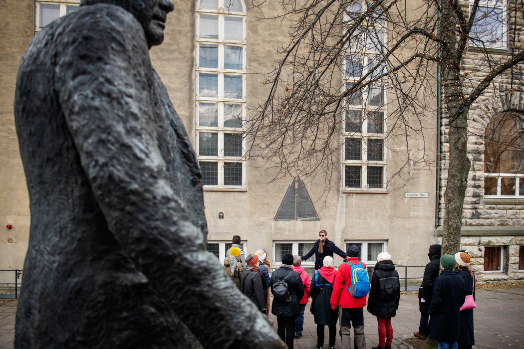I forgrunnen deler av en skulptur av en mann, i bakgrunnen en gruppe mennesker som er på byvandring, midt i gruppen står en mann med utstrakte armer og snakker.