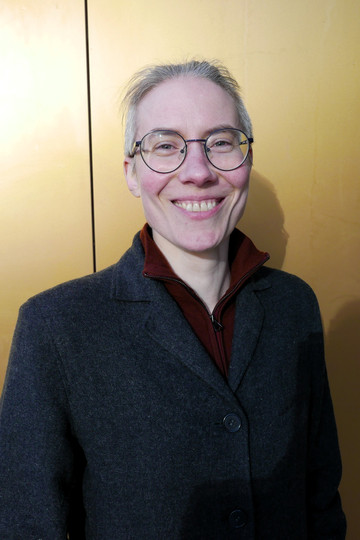 Kvinne med kort hår, briller og grå jakke foran gullfarget bakgrunn.