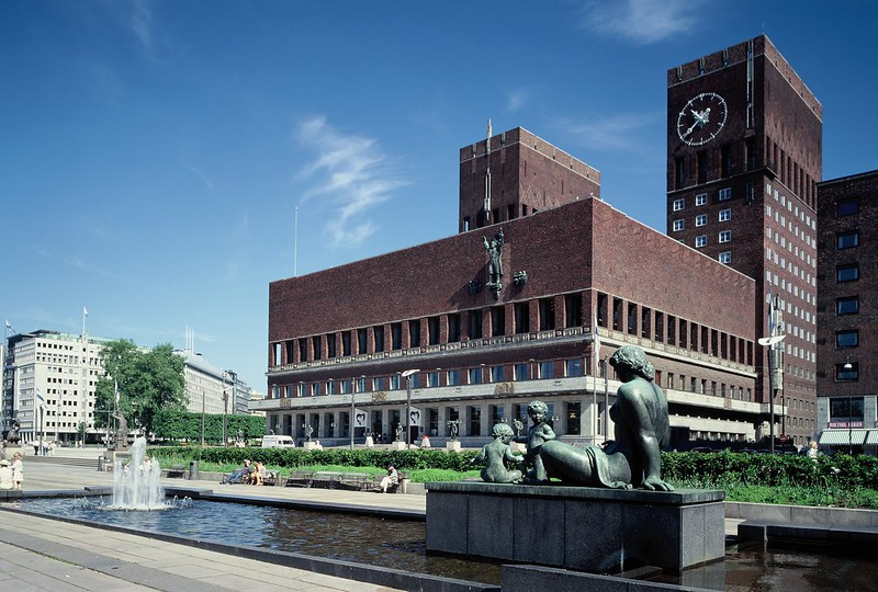 Oslo rådhus sett fra sjøsiden med grønne busker, vannspeil og skulpturer i forgrunnen.
